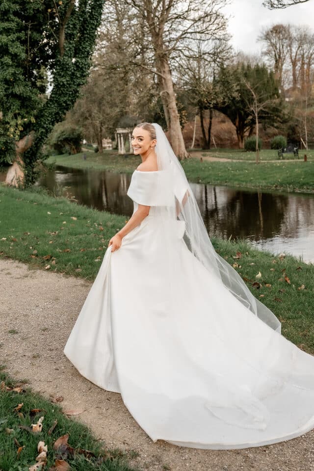 Bride in white gown by a river, wearing a veil and smiling, surrounded by greenery and trees in Bellingham Castle.
