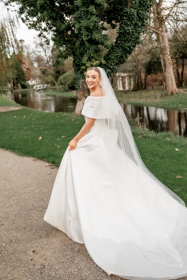 Bride in elegant white gown and veil, smiling by a serene riverside path and lush greenery.