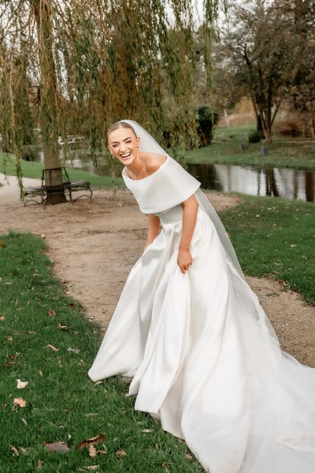 Bride laughing in a white wedding dress with a veil, standing on a garden path by a small pond.