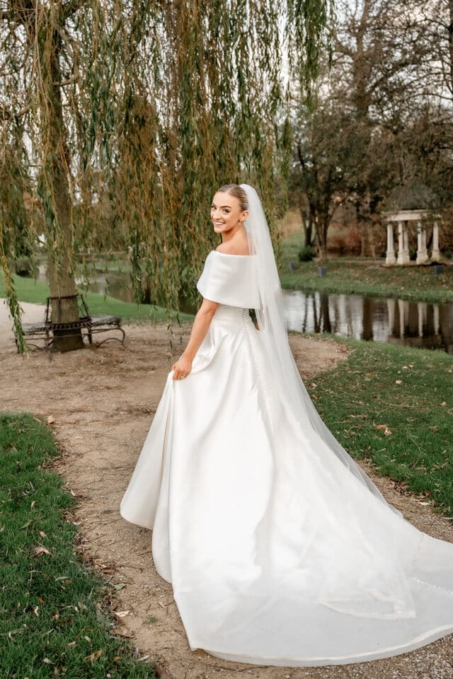 Bride in a white gown posing outdoors near a pond and trees.