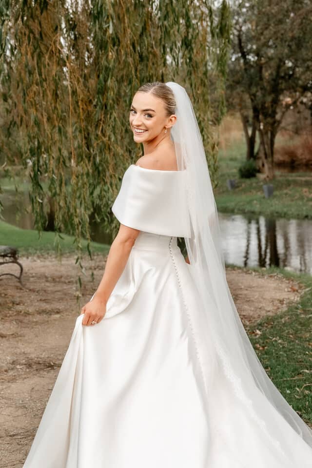 Bride in elegant wedding dress and veil smiling outdoors by greenery.