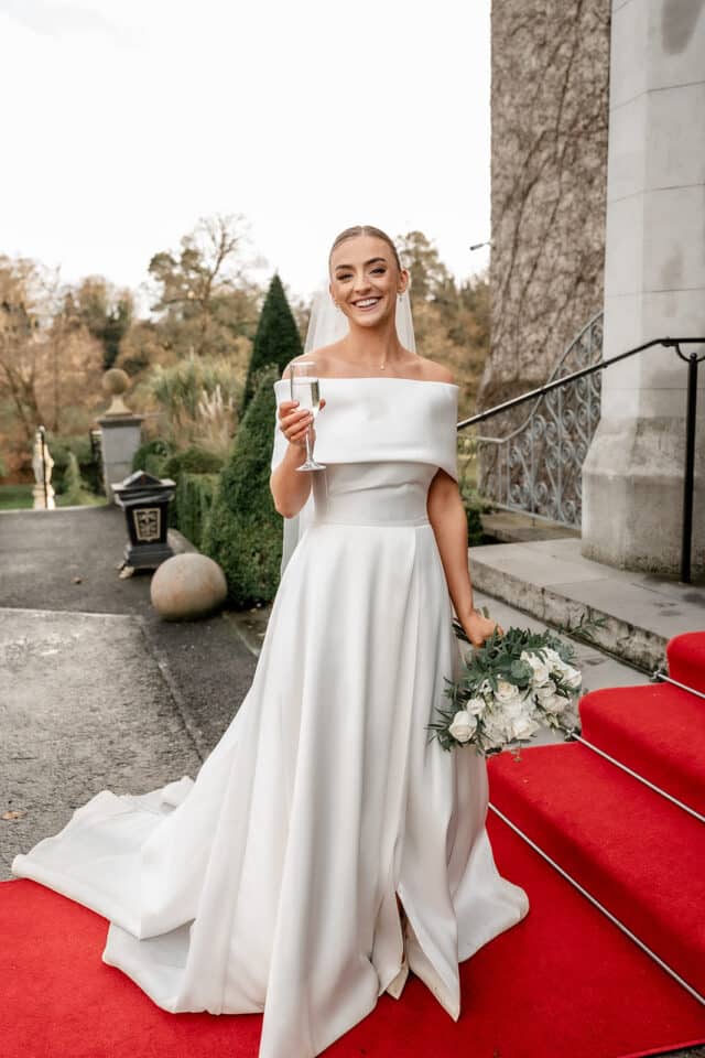 Bride in an elegant white gown holding a bouquet and champagne, standing on red carpet steps at a castle.