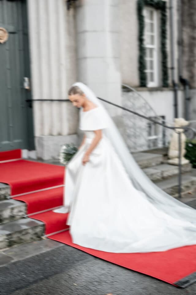 Bride in a white gown walking down red carpet steps at Bellingham Castle.