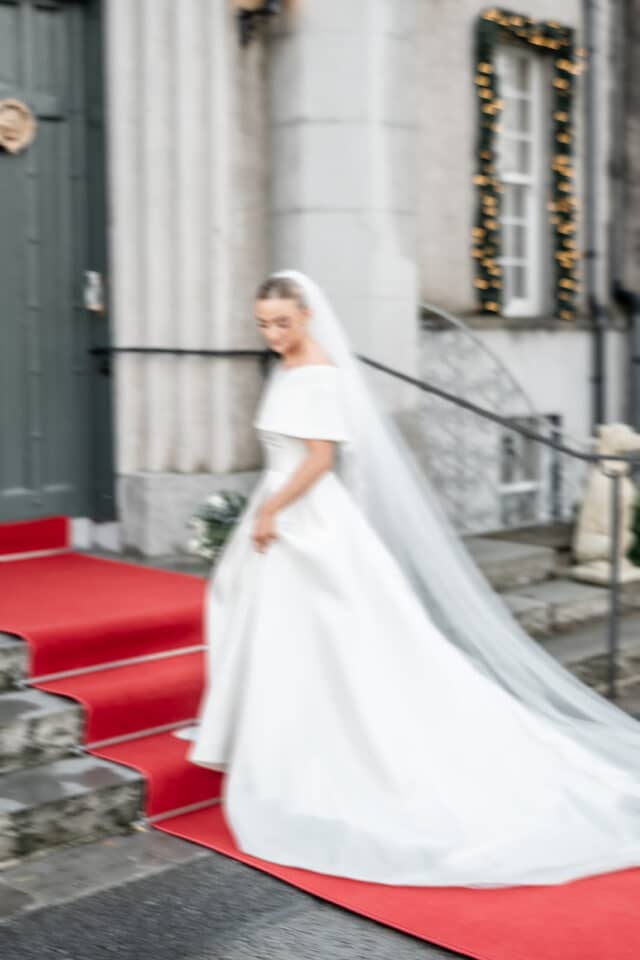 Bride in elegant white gown descending red-carpeted steps outside a castle.
