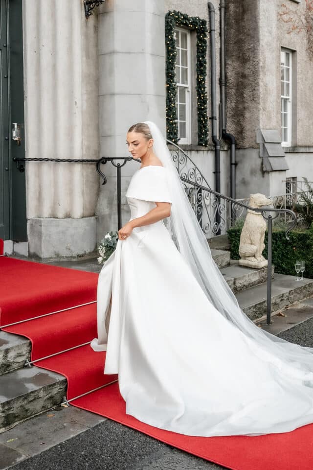 Bride in elegant white gown on red carpet at Bellingham Castle entrance.
