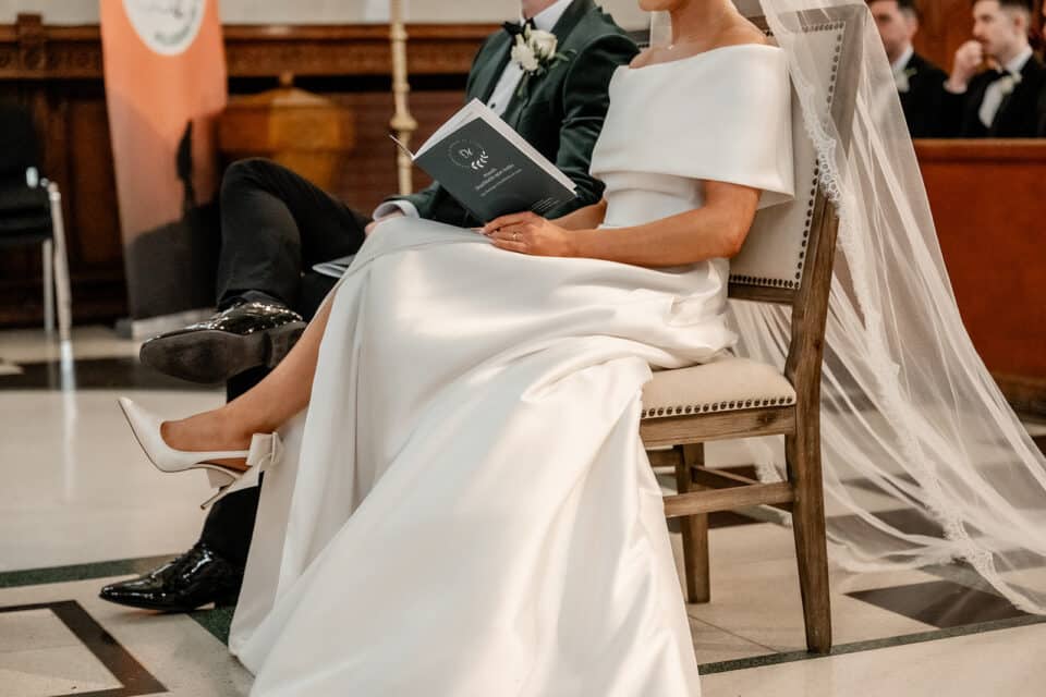 Bride and groom seated at a wedding ceremony, with the bride in a white gown and veil holding a program.