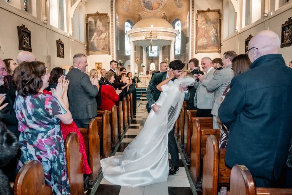 Bride and groom kiss in a church aisle, surrounded by applauding guests.