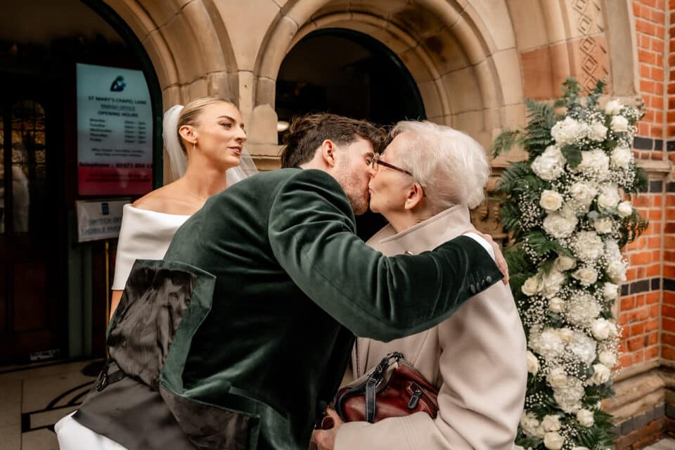 Young couple embraces outside a brick building with floral decorations, man kissing elderly woman.