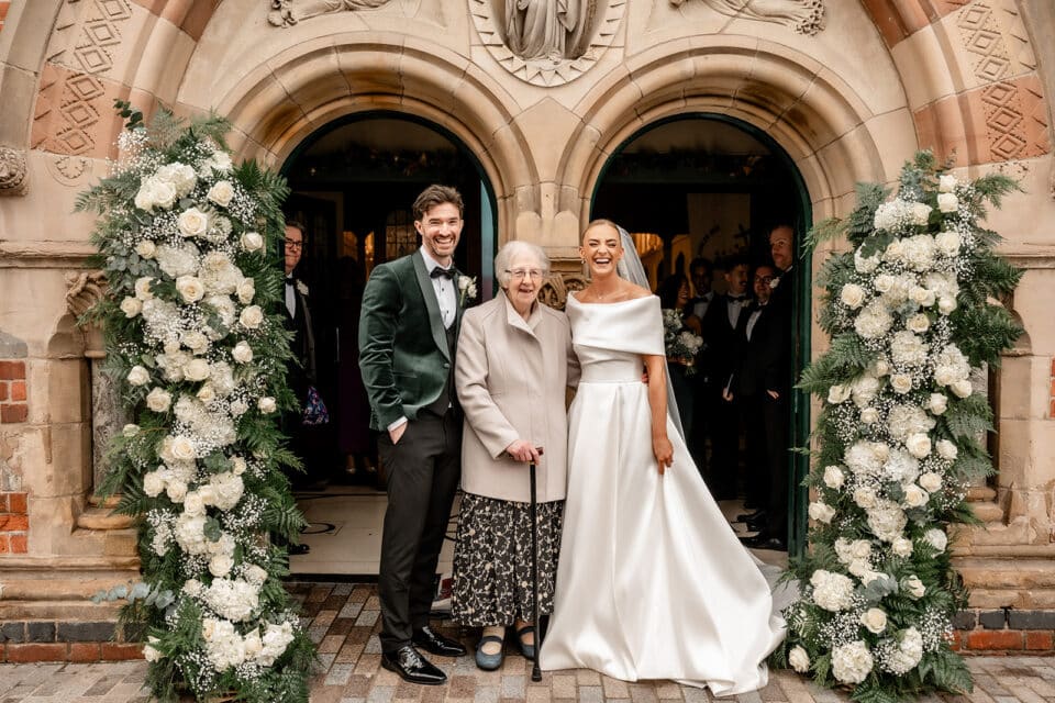 Bride and groom posing with an elderly woman at a flower-adorned stone entrance.