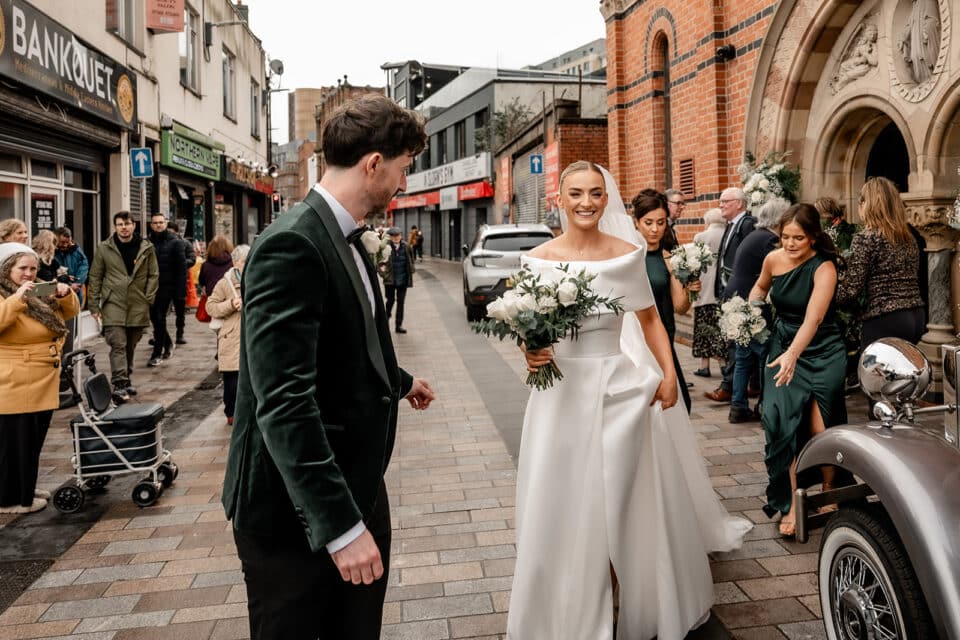 Bride and groom smiling with bridesmaids holding bouquets outside a building, with onlookers in the background.
