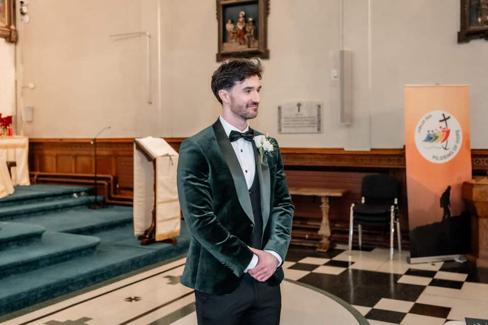 Groom in a green velvet suit waits at the altar in a church setting.