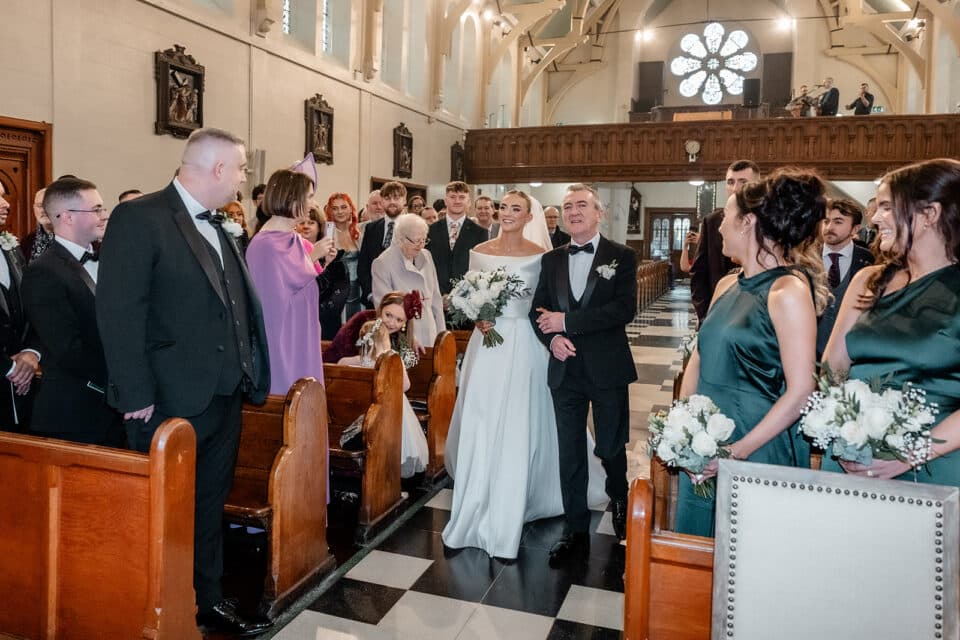 Bride walking down the aisle with her father in a church, surrounded by guests and bridesmaids.