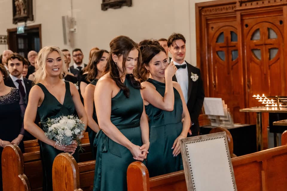Bridesmaids in green dresses and guests in a church, smiling and emotional during a wedding ceremony.