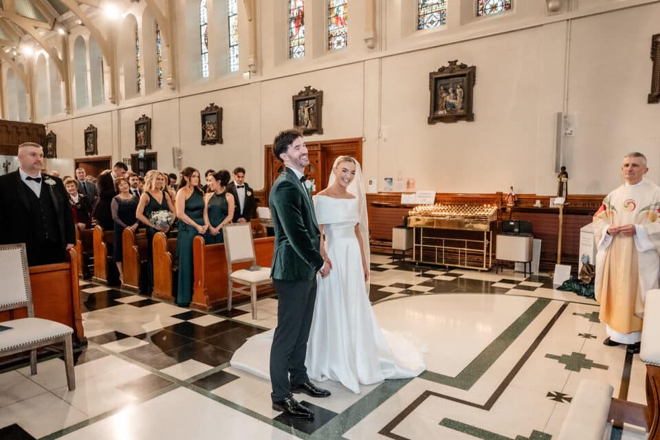 Bride and groom smile during their wedding ceremony in a church with guests and a priest present.