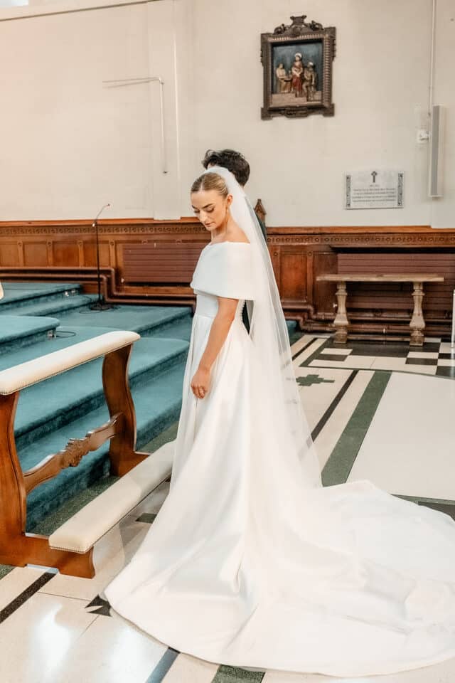 Bride in elegant white gown and veil standing in a church interior near an altar.
