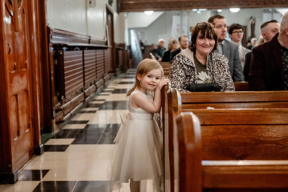 Young girl in a white dress smiling in a church pew, surrounded by seated guests.