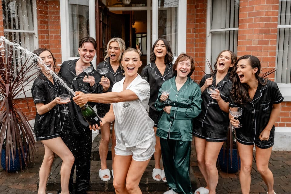 Group celebrating with champagne, wearing matching pajamas outside a brick building.