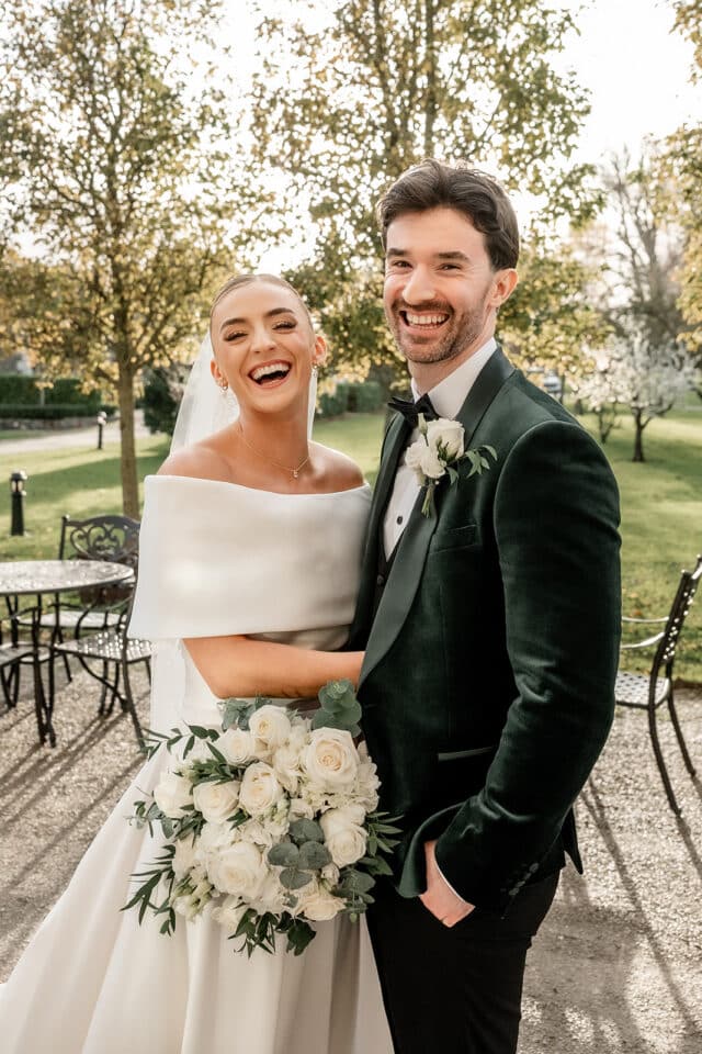 Bride and groom smiling outdoors at Bellingham Castle, with trees and greenery in the background.