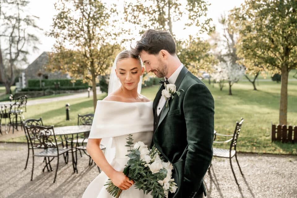 Bride and groom embracing outdoors at Bellingham Castle, surrounded by greenery, holding a white rose bouquet.