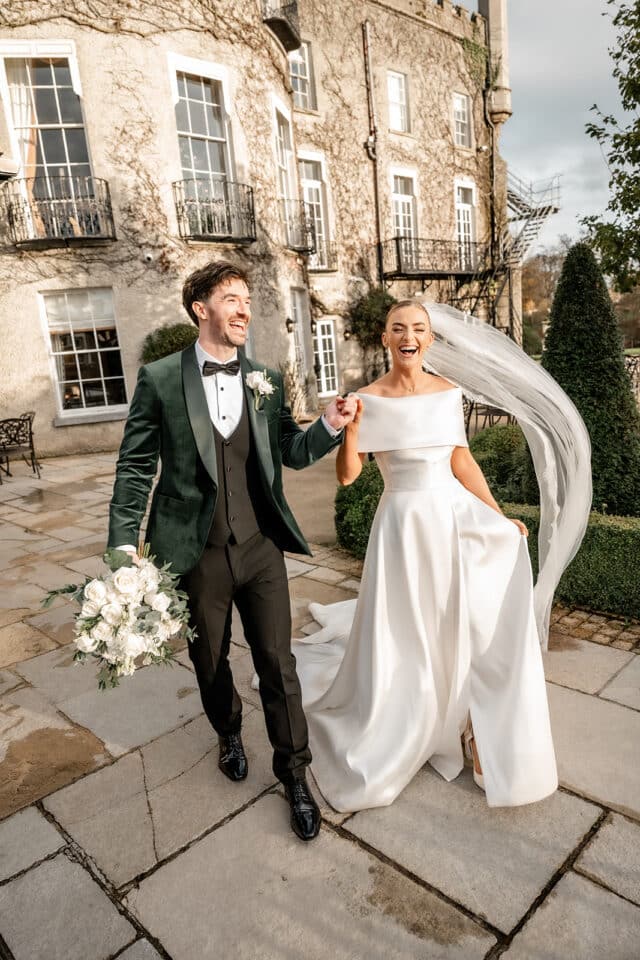 Bride and groom joyfully walking outside Bellingham Castle, holding hands and a bouquet, with elegant attire and veil.