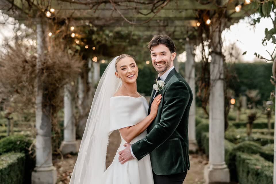 Bride and groom smiling under pergola at Bellingham Castle garden.