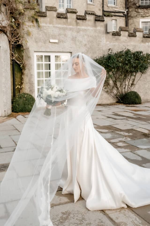 Bride in an elegant wedding dress holding a bouquet, draped in a long veil outside a stone castle.