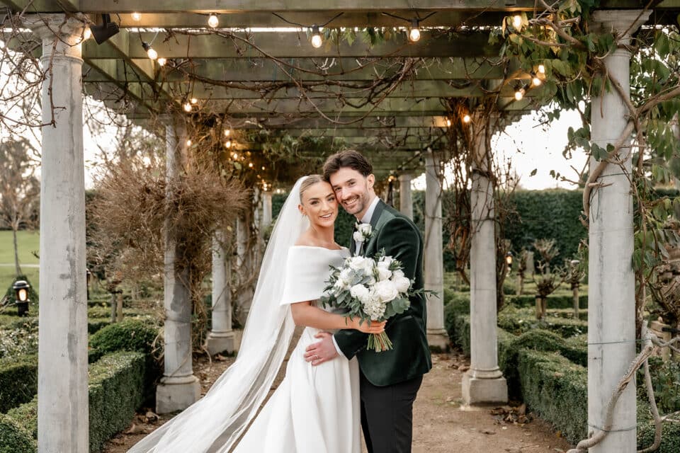 Bride and groom smiling under pergola at Bellingham Castle garden.