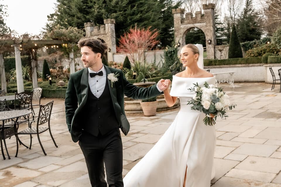 Bride and groom holding hands and walking at Bellingham Castle, with the bride in a white dress and bouquet.