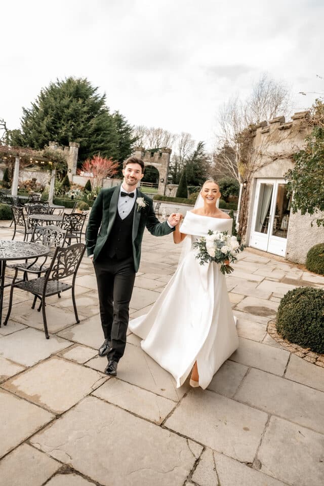 Bride and groom walking joyfully at Bellingham Castle courtyard, surrounded by gardens.