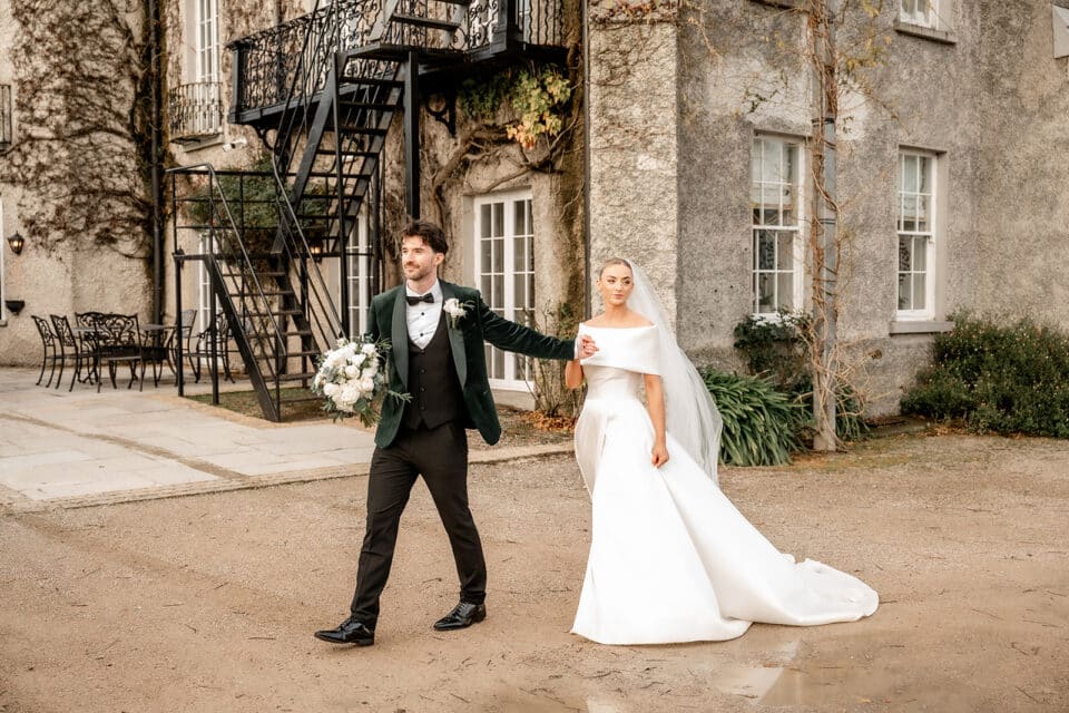 Bride and groom walk holding hands at Bellingham Castle, with the groom holding a bouquet.