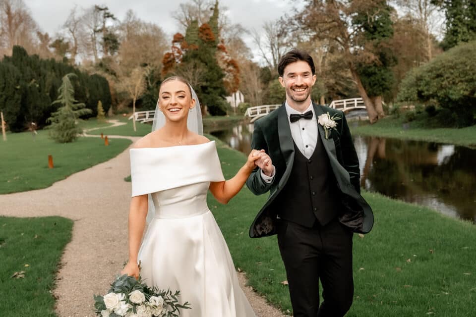Bride and groom holding hands, walking on a garden path near a pond, Bellingham Castle.