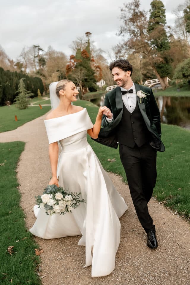 Bride and groom walking hand-in-hand on a path, bride holding a bouquet of white flowers.