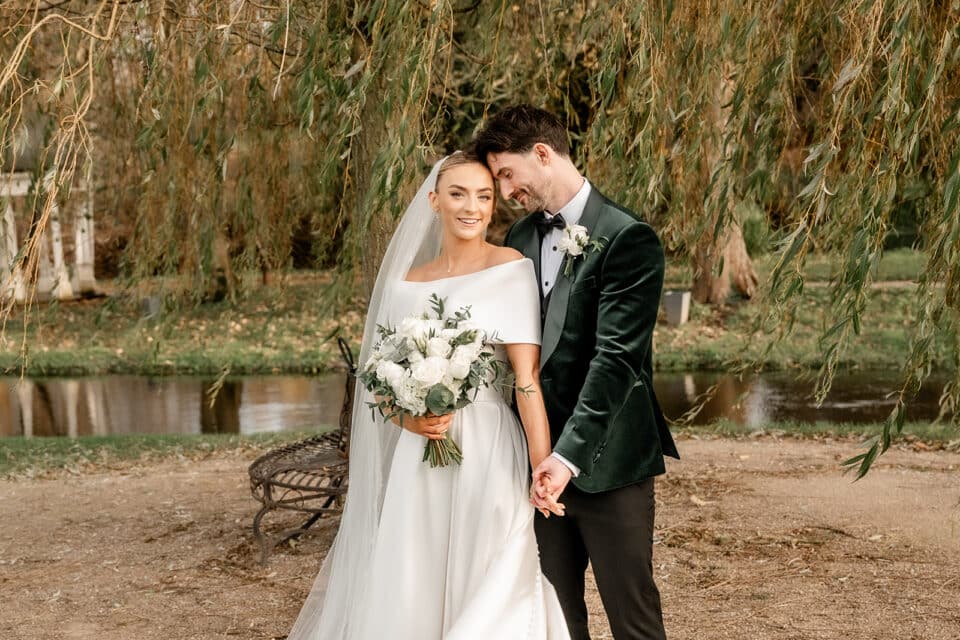 Bride and groom posing happily under a tree near a pond at Bellingham Castle.