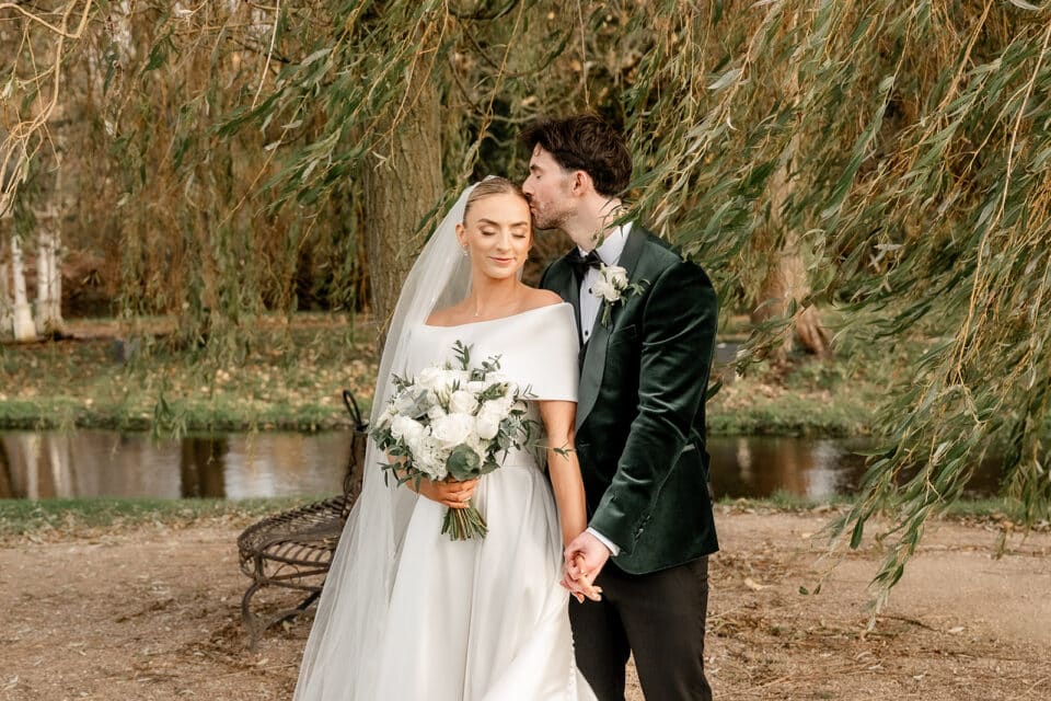 Bride and groom at Bellingham Castle, with groom kissing bride's head, holding hands near a pond under willow branches.