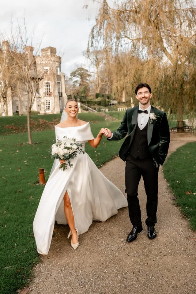 Bride and groom walking on a path at Bellingham Castle, bride in white gown holding bouquet, groom in dark suit.