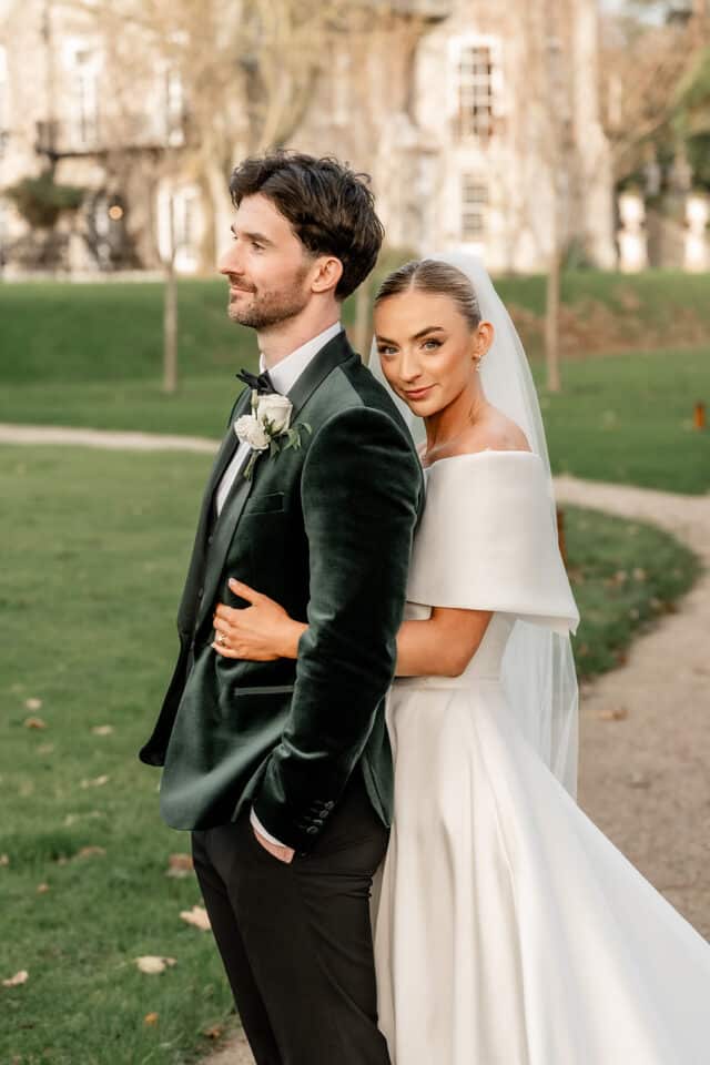 Bride in white gown hugs groom in dark suit outside Bellingham Castle.