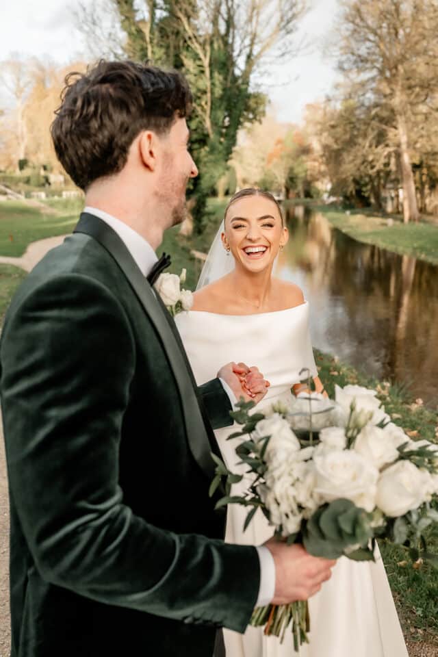 Bride and groom smiling by a riverside, holding hands and bouquets, with autumn trees in the background at Bellingham Castle.