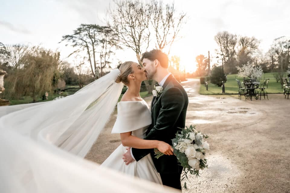 Bride and groom kissing outdoors at sunset, with the bride holding a bouquet and wearing a long veil.