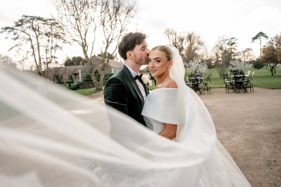 Bride and groom embrace outdoors at Bellingham Castle, with veil flowing and elegant attire.