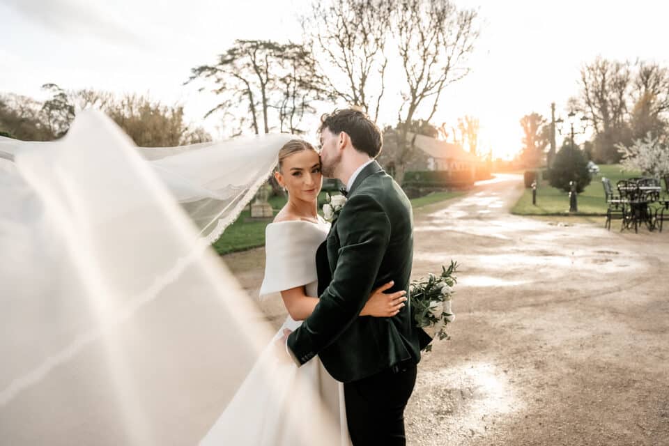 Bride and groom embrace outdoors with flowing veil at sunset.