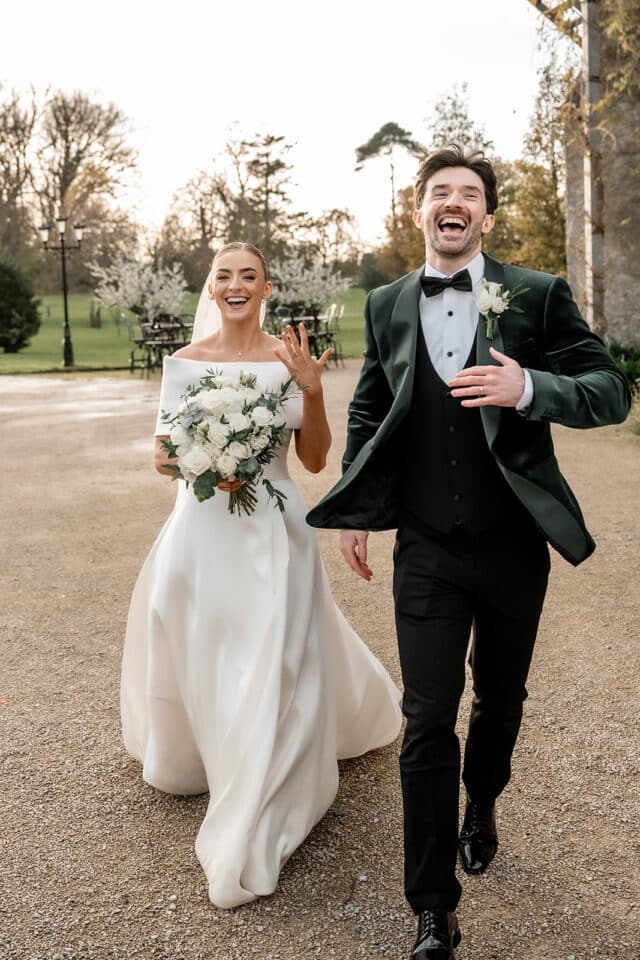 Bride and groom joyfully walking outside a castle, bride holding bouquet, and groom in a tuxedo.