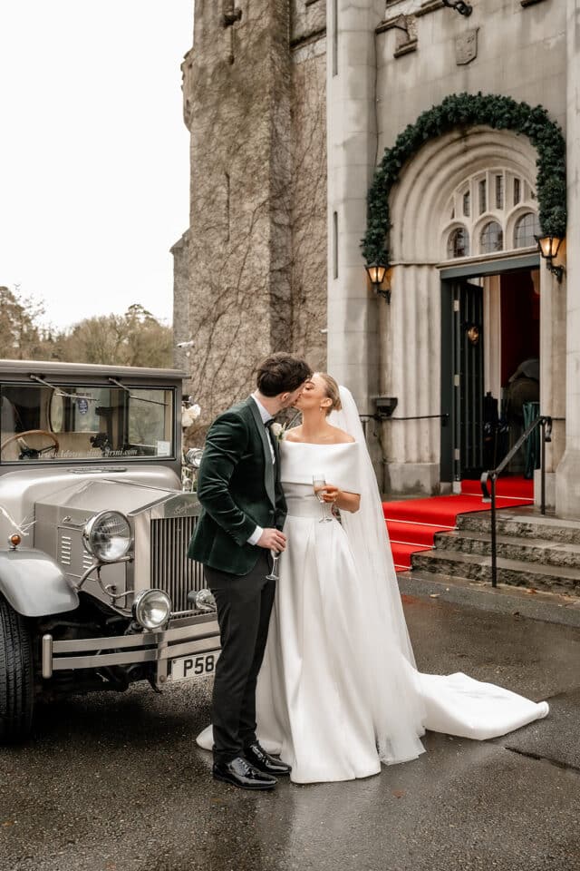 Bride and groom kiss outside Bellingham Castle, holding champagne glasses beside a vintage car.