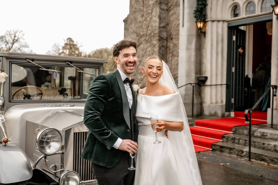 Bride and groom smiling beside vintage car at Bellingham Castle.