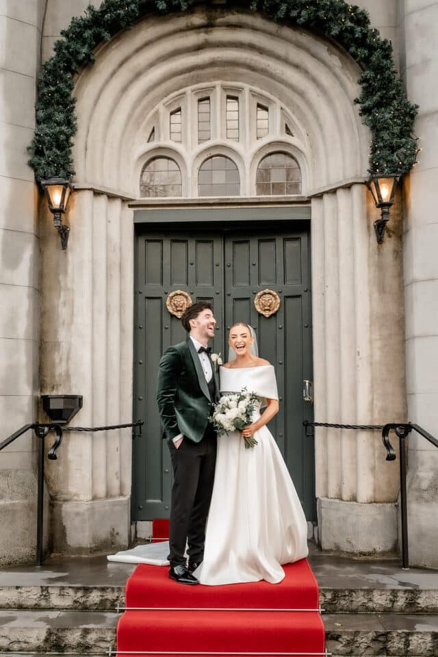 Bride and groom smiling on red carpet at Bellingham Castle entrance.