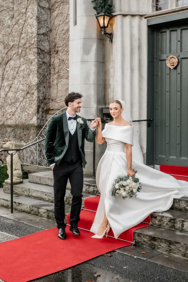 Bride and groom holding hands on red carpet, bride in white gown, groom in dark suit, at castle entrance.