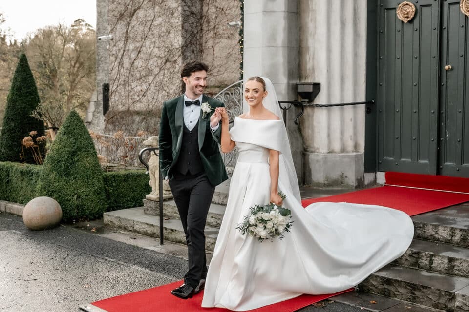 Bride and groom smiling, walking down a red carpet at Bellingham Castle.