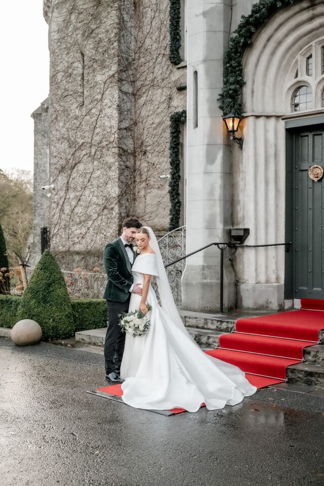 Bride and groom standing on red carpet at Bellingham Castle entrance, embracing in wedding attire.
