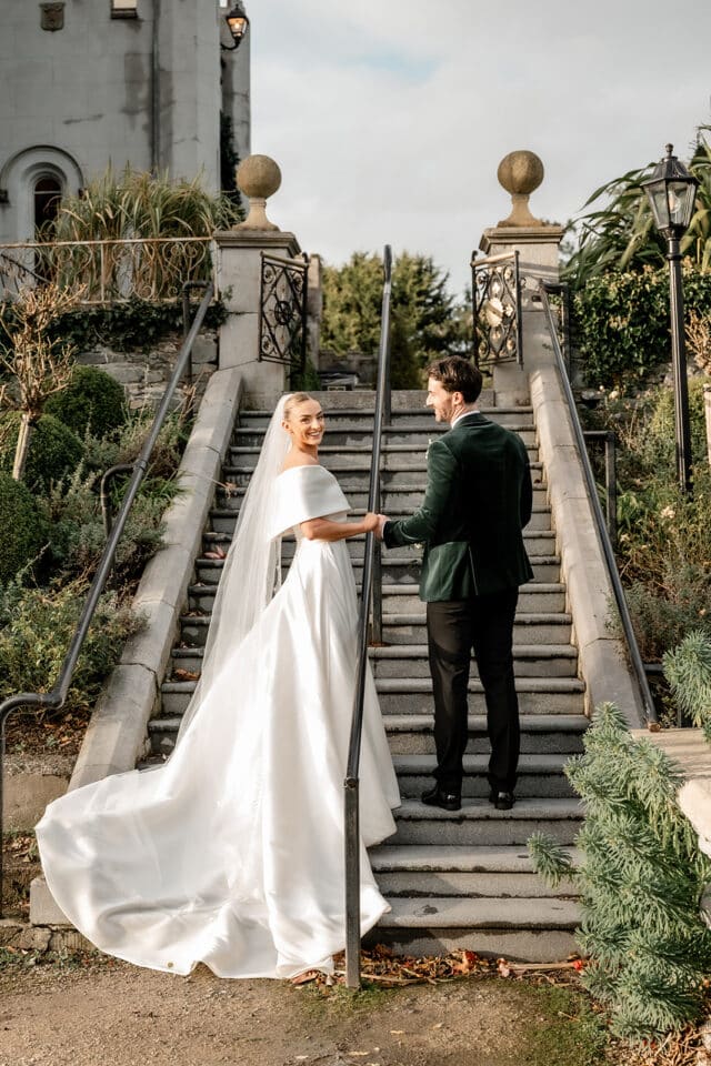Bride and groom holding hands on castle steps, surrounded by greenery.