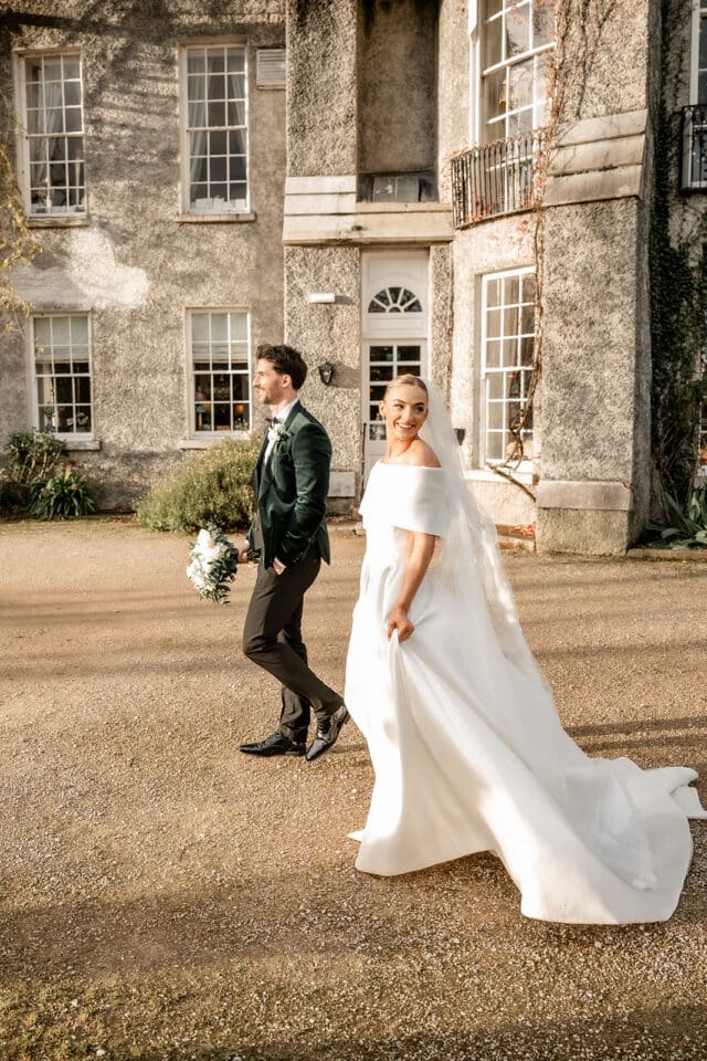 Bride and groom walking outside Bellingham Castle, bride in a white dress and groom in a dark suit holding flowers.