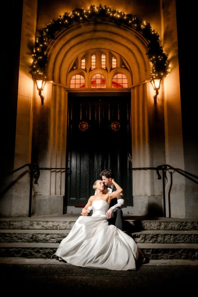Bride and groom sitting on stairs at Bellingham Castle, illuminated by warm lights, outside a decorated entrance.